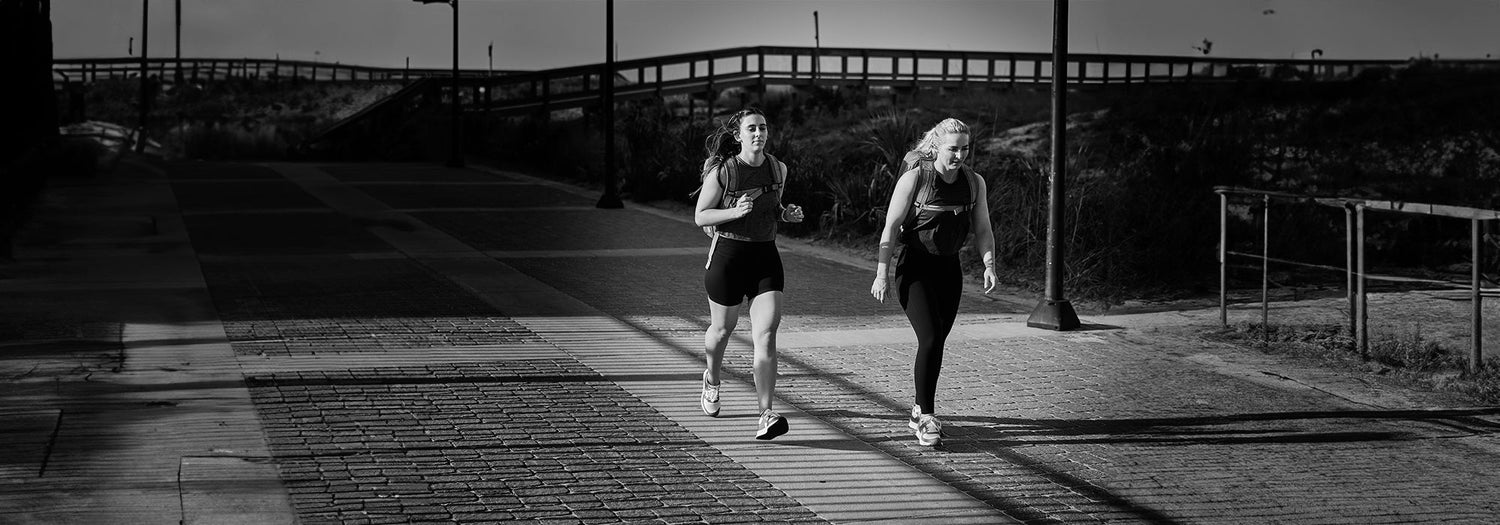 Two people jogging on a paved path near a wooden fence with a sunset backdrop.