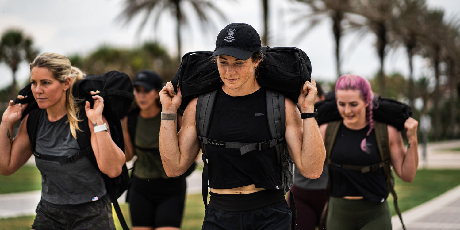 Four women exercising outdoors, carrying weighted backpacks on their shoulders, with grassy background and palm trees.