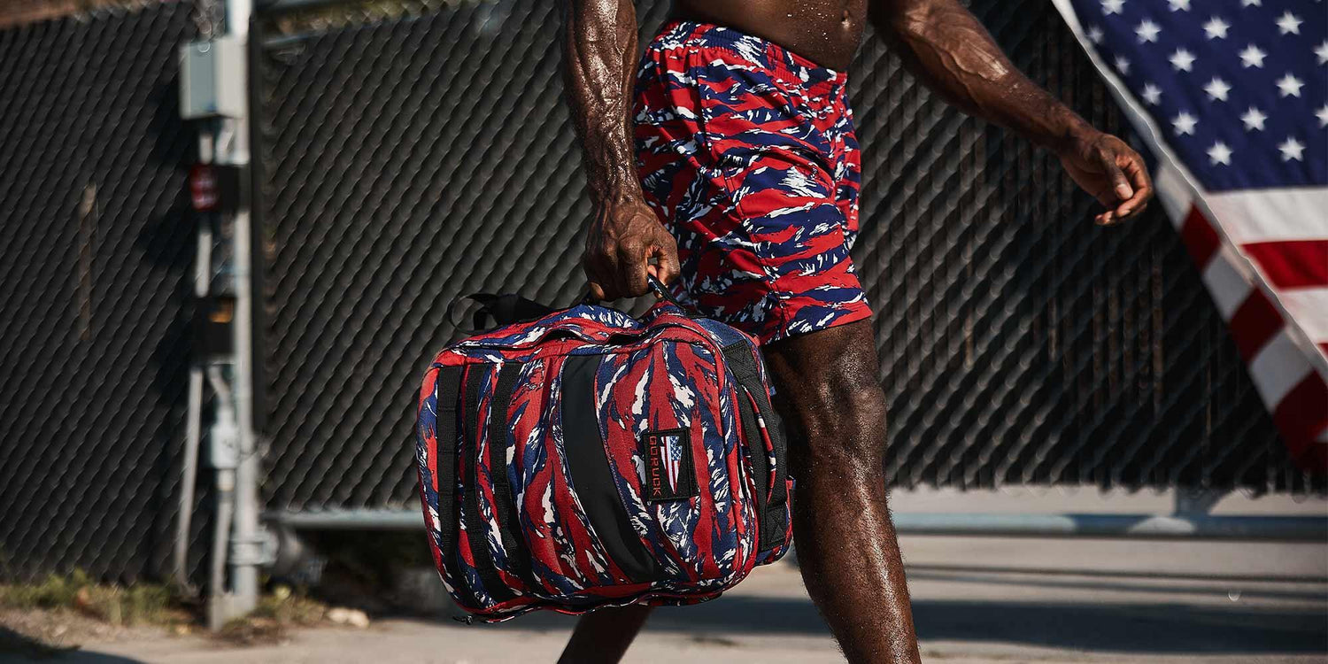 Fit man carrying red, white, and blue tiger stripe GORUCK bag wearing matching shorts with American flag in background
