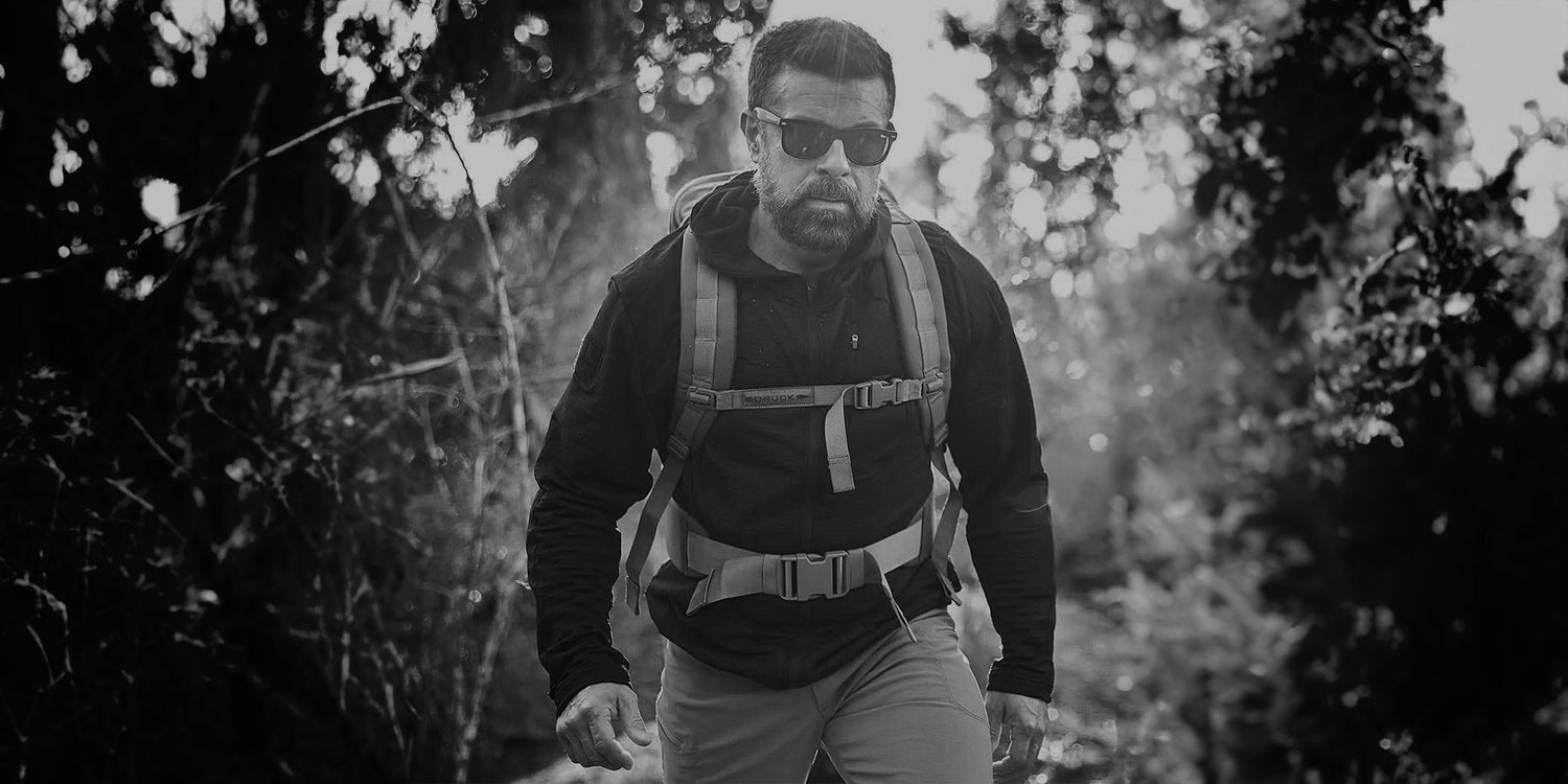 Man wearing GORUCK tactical backpack and outdoor apparel hiking through sunlit forest trail