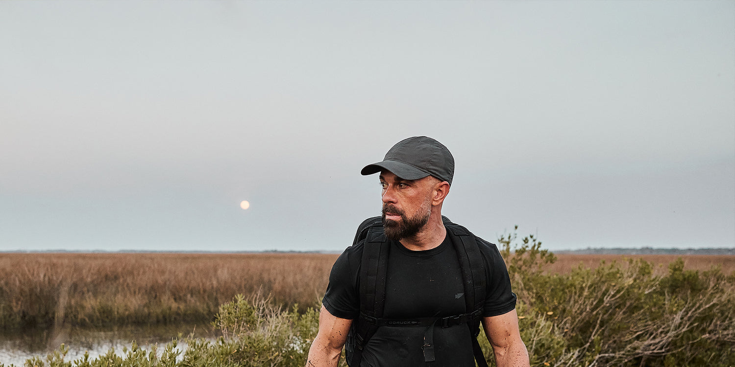 Man wearing black GORUCK gear and cap standing in outdoor marshland during early evening