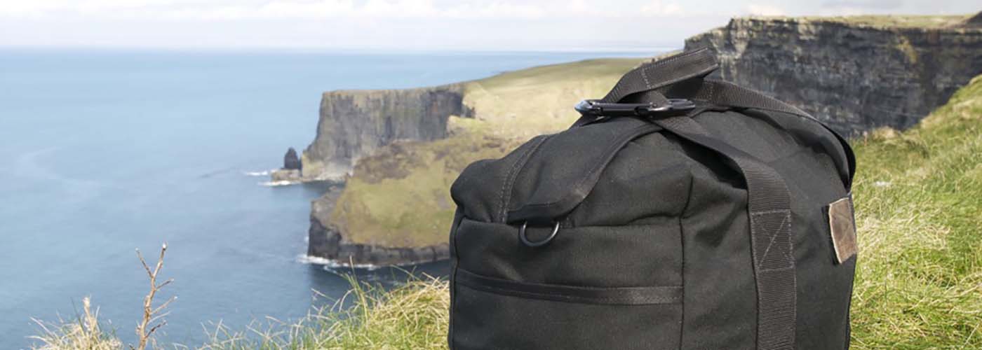 Black GORUCK tactical bag on grassy cliff edge overlooking ocean and rugged coastline