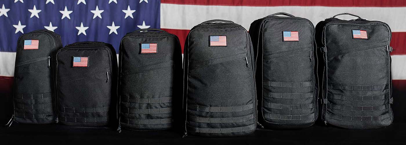 Six black GORUCK Cordura backpacks with American flag patches lined up in front of a large US flag