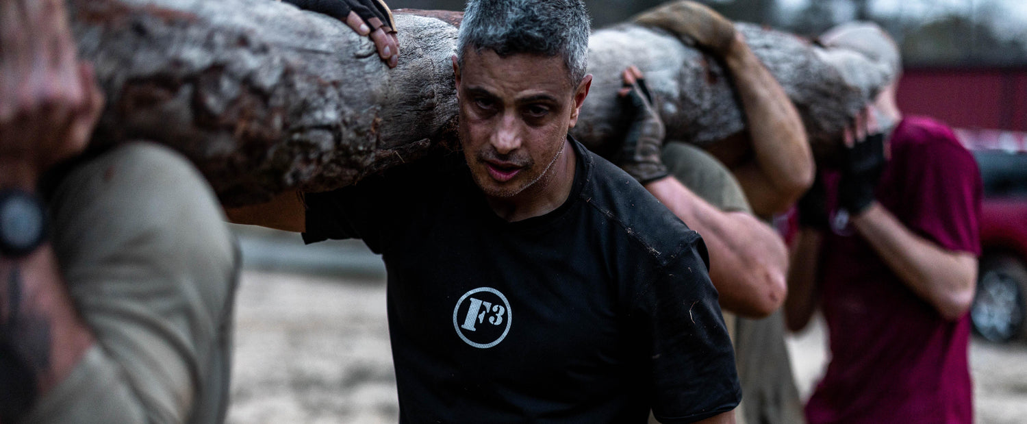 Team rucking challenge with men carrying heavy log outdoors in GORUCK training gear