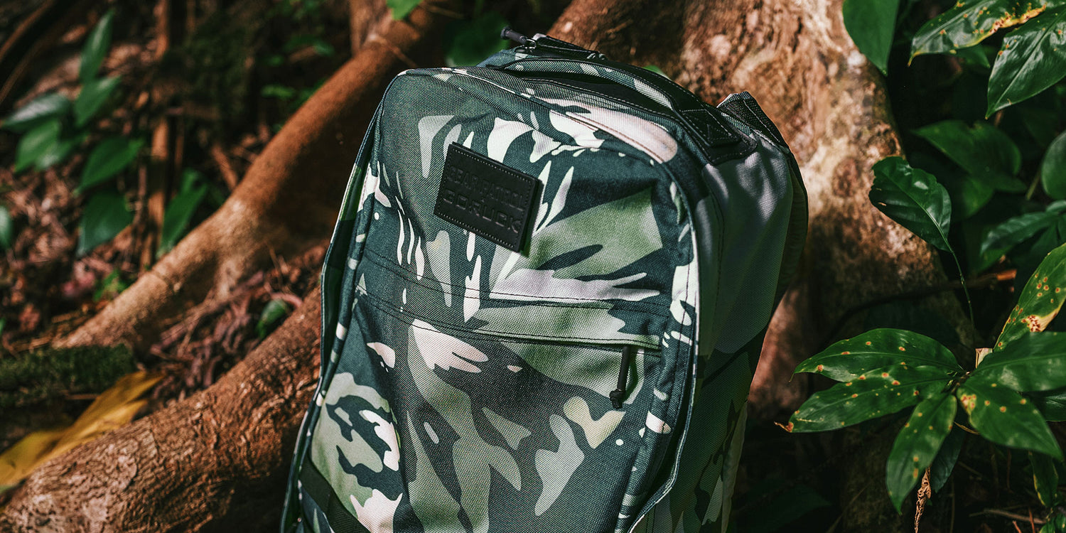 Camouflage backpack leaning against a tree trunk in a lush green forest setting.