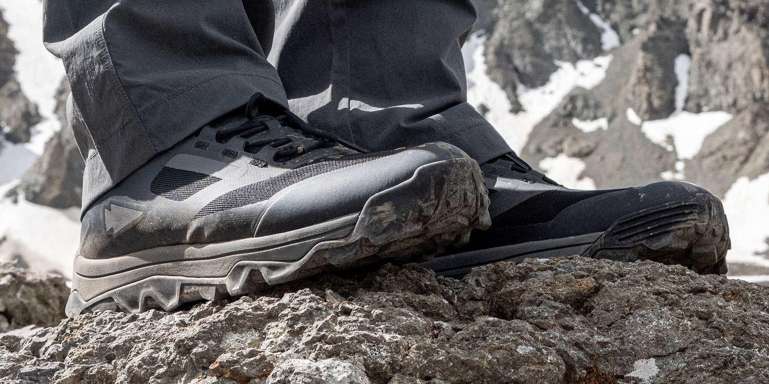 Close-up of person wearing rugged hiking boots standing on rocky terrain with mountains in the background.