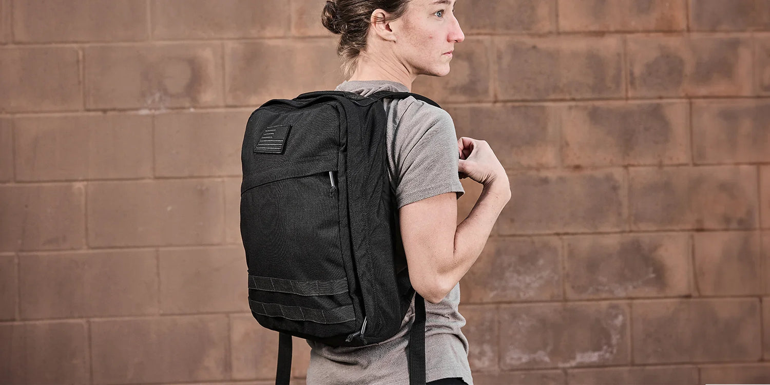 Woman wearing GORUCK black rucksack backpack against brick wall, rugged outdoor gear