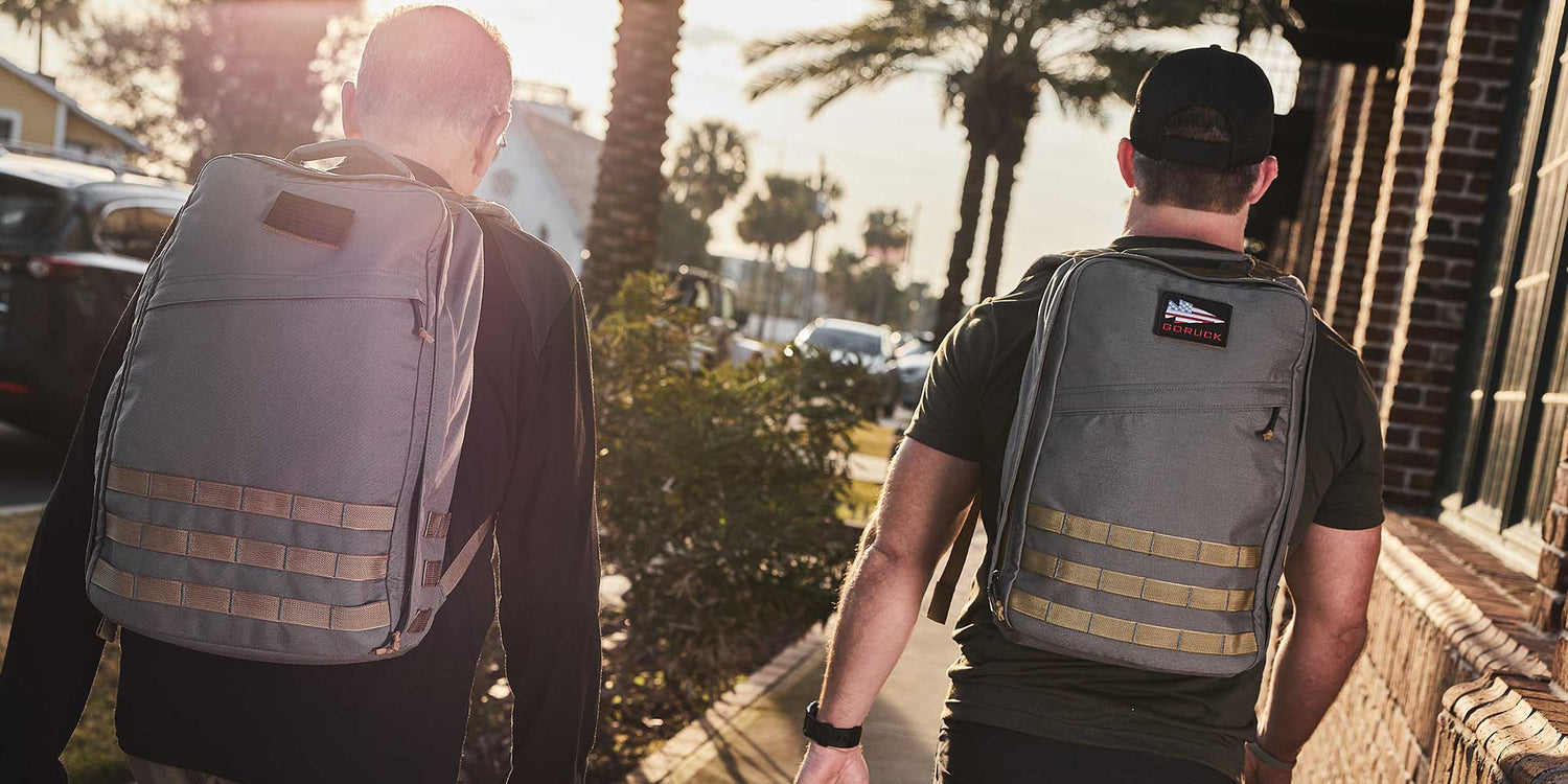 Two men walking outdoors in sunlight wearing durable grey GORUCK backpacks on a palm tree-lined street