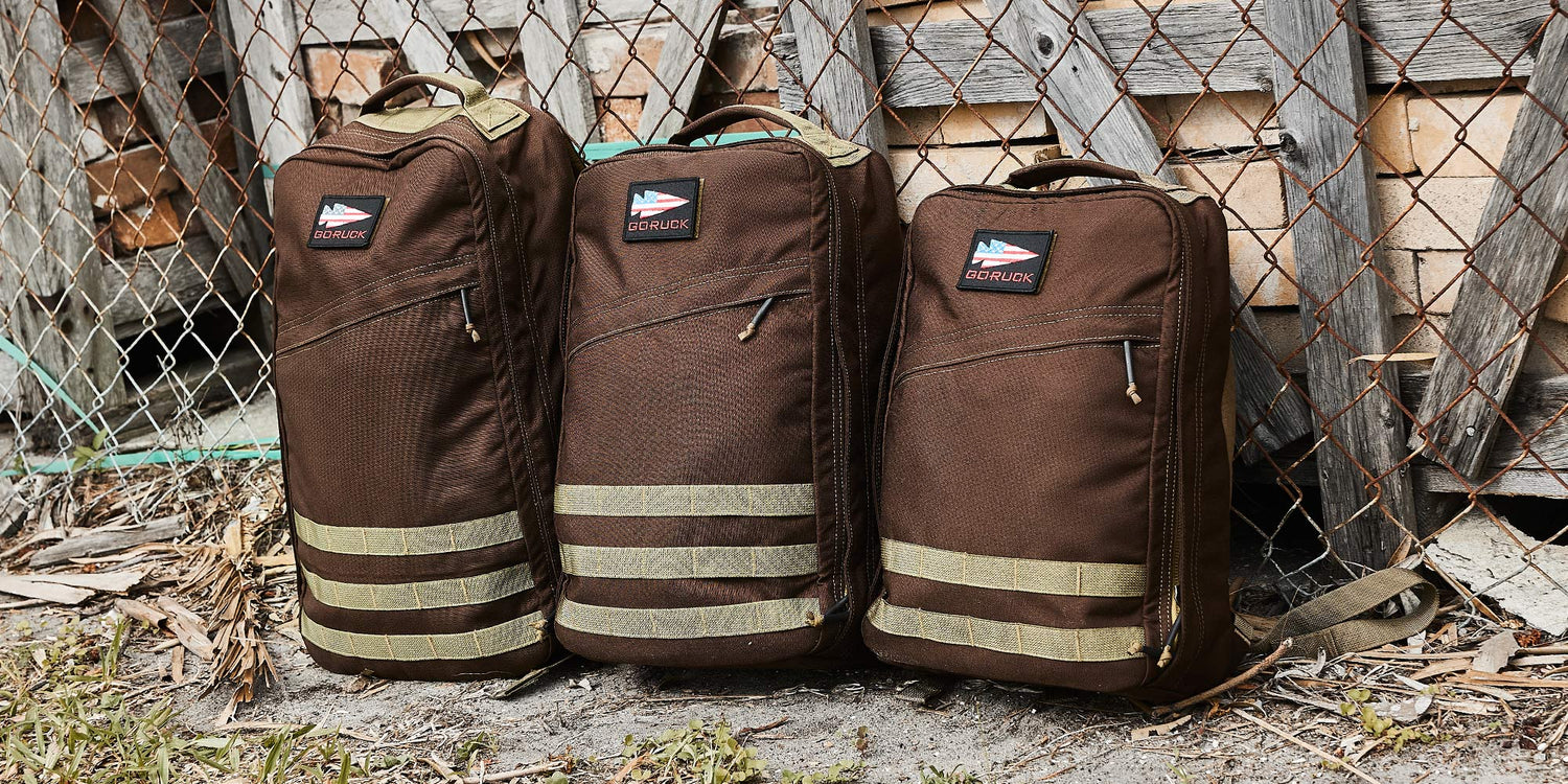Three brown GORUCK rucksacks with beige straps against a rustic, weathered wooden fence