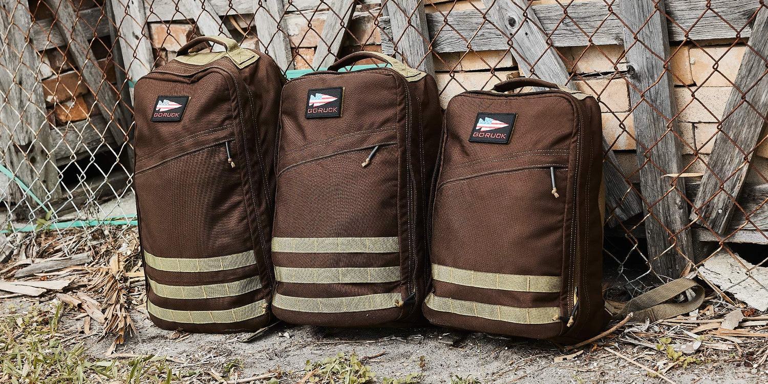 Three brown GORUCK rucksacks with beige straps against a rustic, weathered wooden fence
