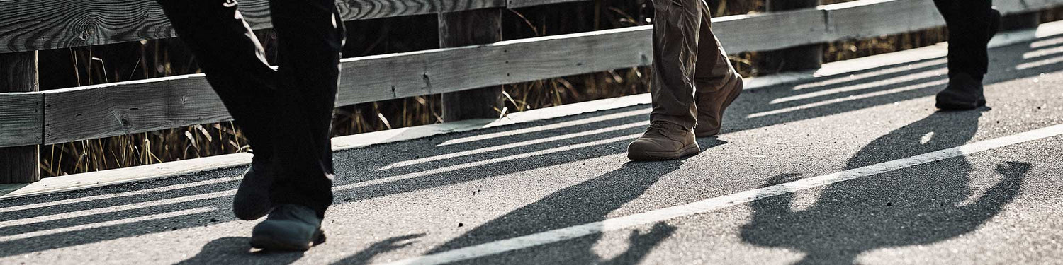 Close-up of three people walking on a sunlit path next to a wooden fence, casting long shadows.