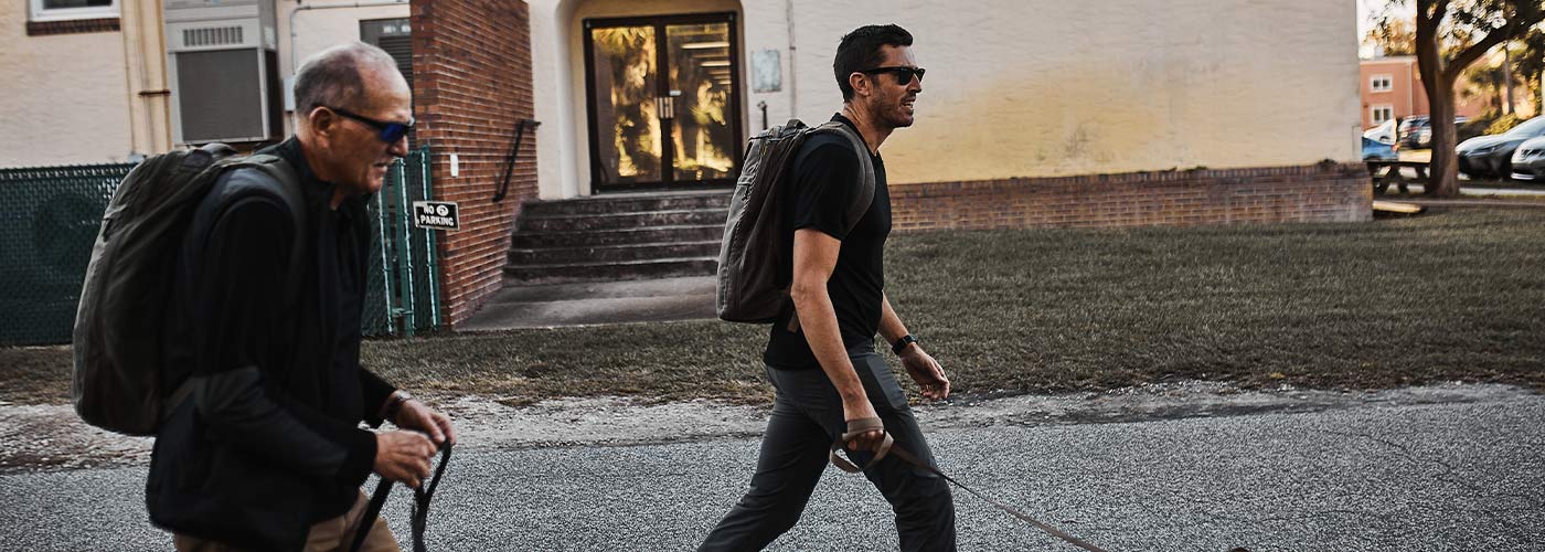 Two men wearing sunglasses walk outside along a street, each carrying backpacks and using walking sticks.