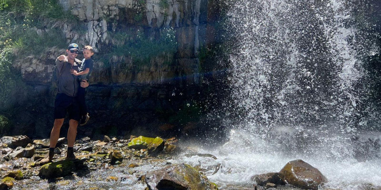 Man and child outdoors by rocky waterfall, wearing hiking gear and boots, GORUCK rucking scene