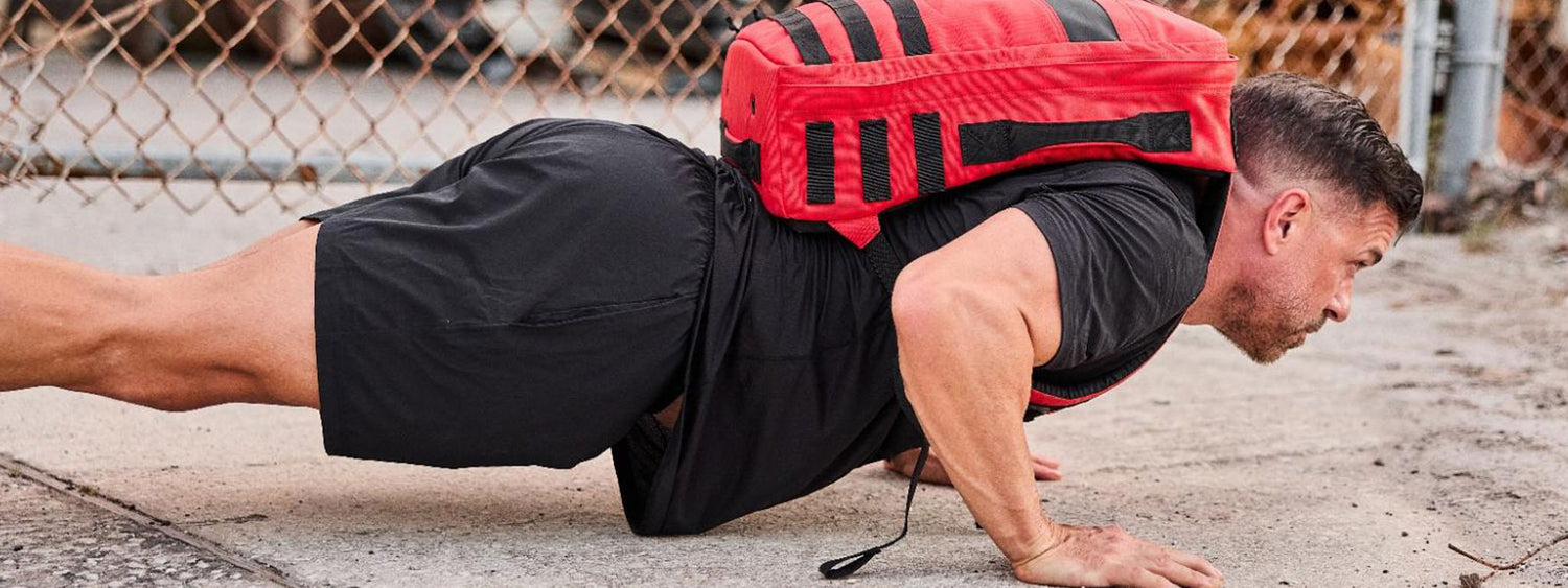 Man doing a push-up on concrete while wearing a red weighted backpack and black workout clothes.