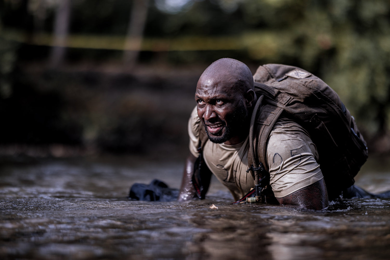 Person in a muddy creek, wearing a backpack and camouflage clothing, appears focused and determined.