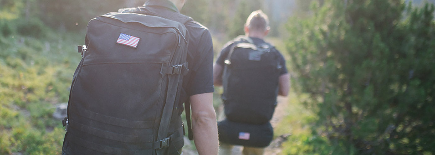 Two hikers wearing sturdy black GORUCK travel rucksacks with American flag patches walking on a sunlit forest trail