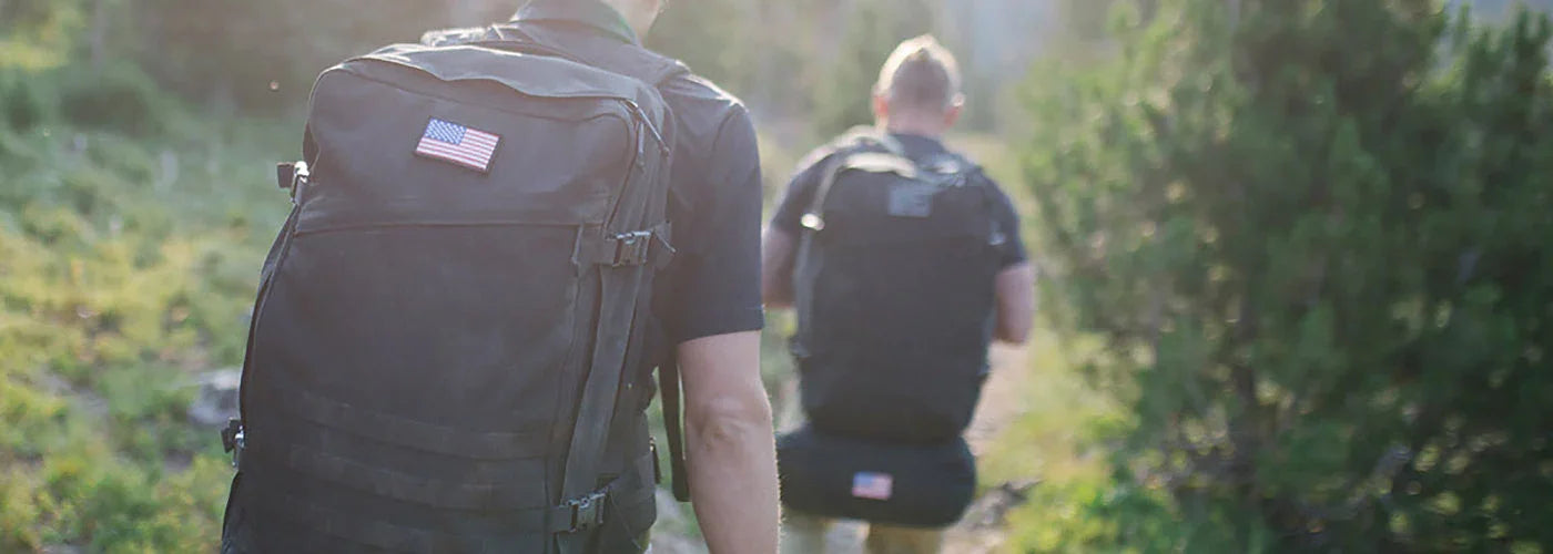 Two hikers wearing sturdy black GORUCK travel rucksacks with American flag patches walking on a sunlit forest trail