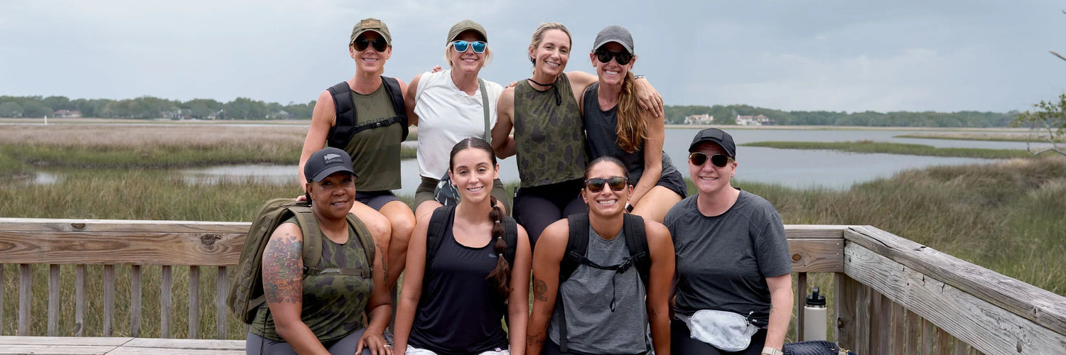 Group of eight women wearing GORUCK outdoor gear posing on wooden deck with marshland and water in background