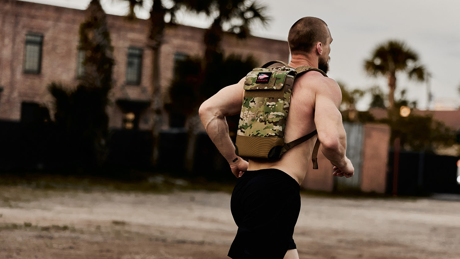 Shirtless man running outdoors with a camouflage backpack, buildings and palm trees in the background.
