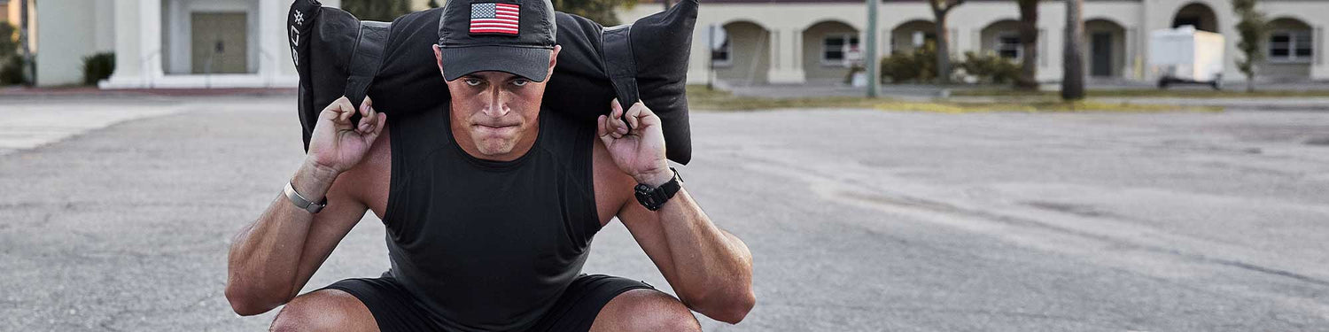 Man in athletic gear squatting outdoors, wearing a cap and lifting a sandbag across his shoulders.