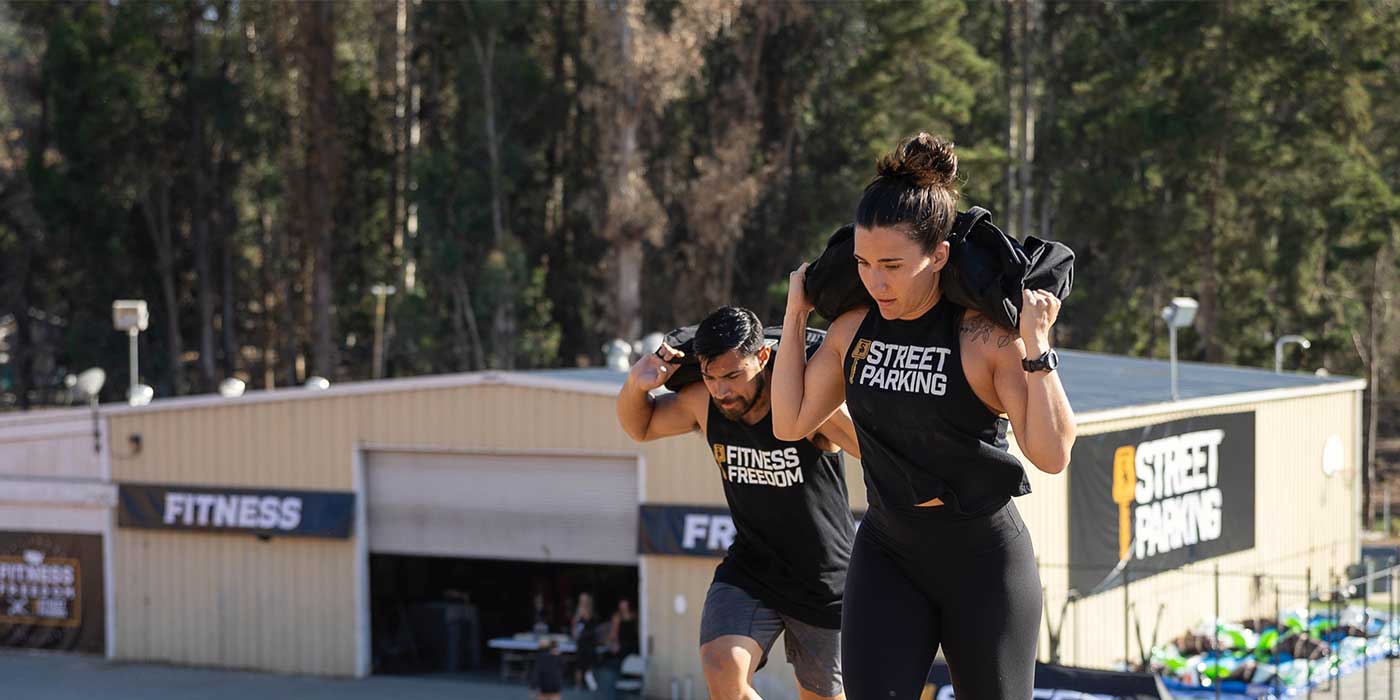Two people carry sandbags uphill outdoors near a fitness center, both wearing athletic
