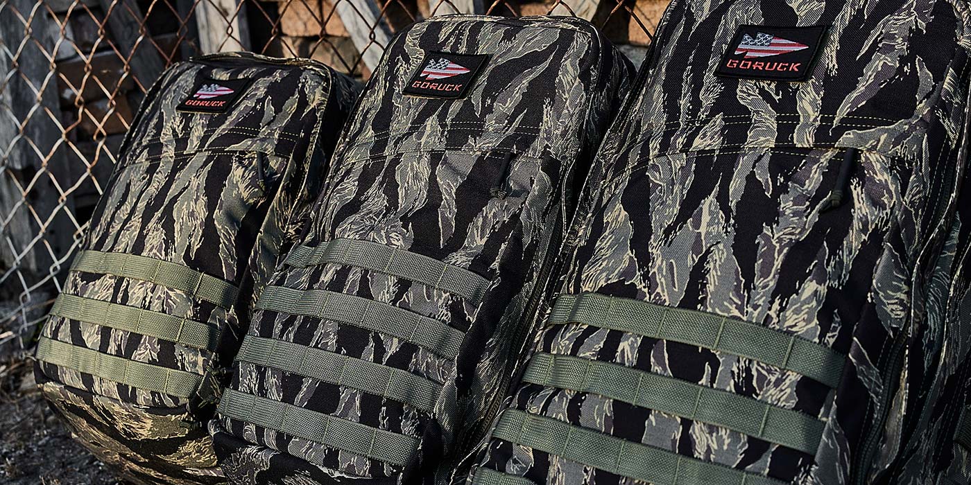 Three GORUCK branded tactical rucking backpacks in tiger stripe camouflage pattern leaning against a chain-link fence