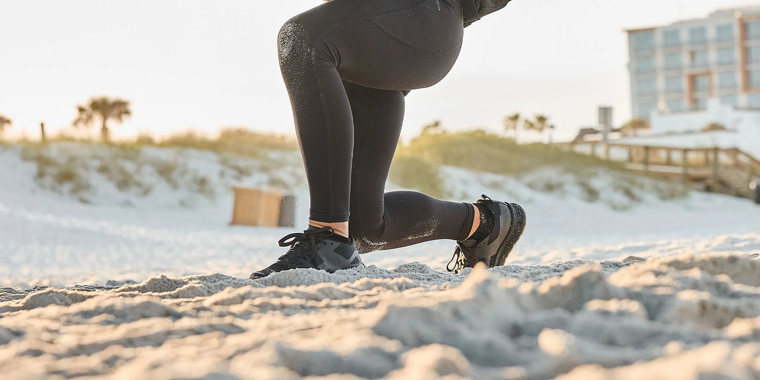 Person in black leggings and sneakers doing lunges on a sandy beach with buildings in the background.