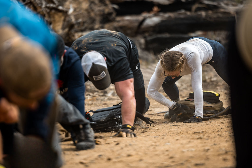 Two people kneel on a sandy ground, leaning forward with backpacks, appearing focused and exerted.