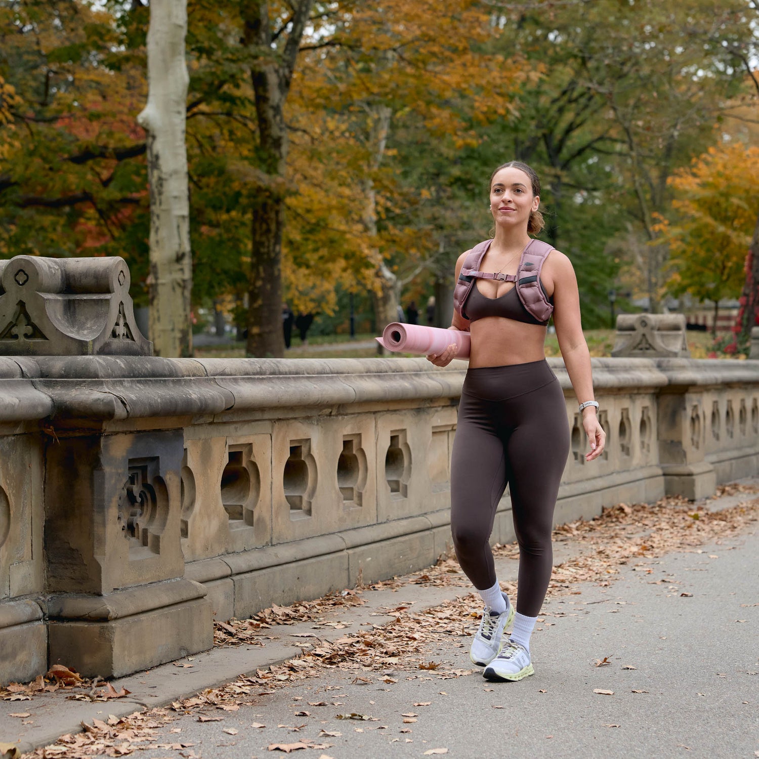 Woman in athletic wear holding a pink yoga mat and the Spy Ruck | Women's Weighted Vest, walking through a park with autumn trees in the background.