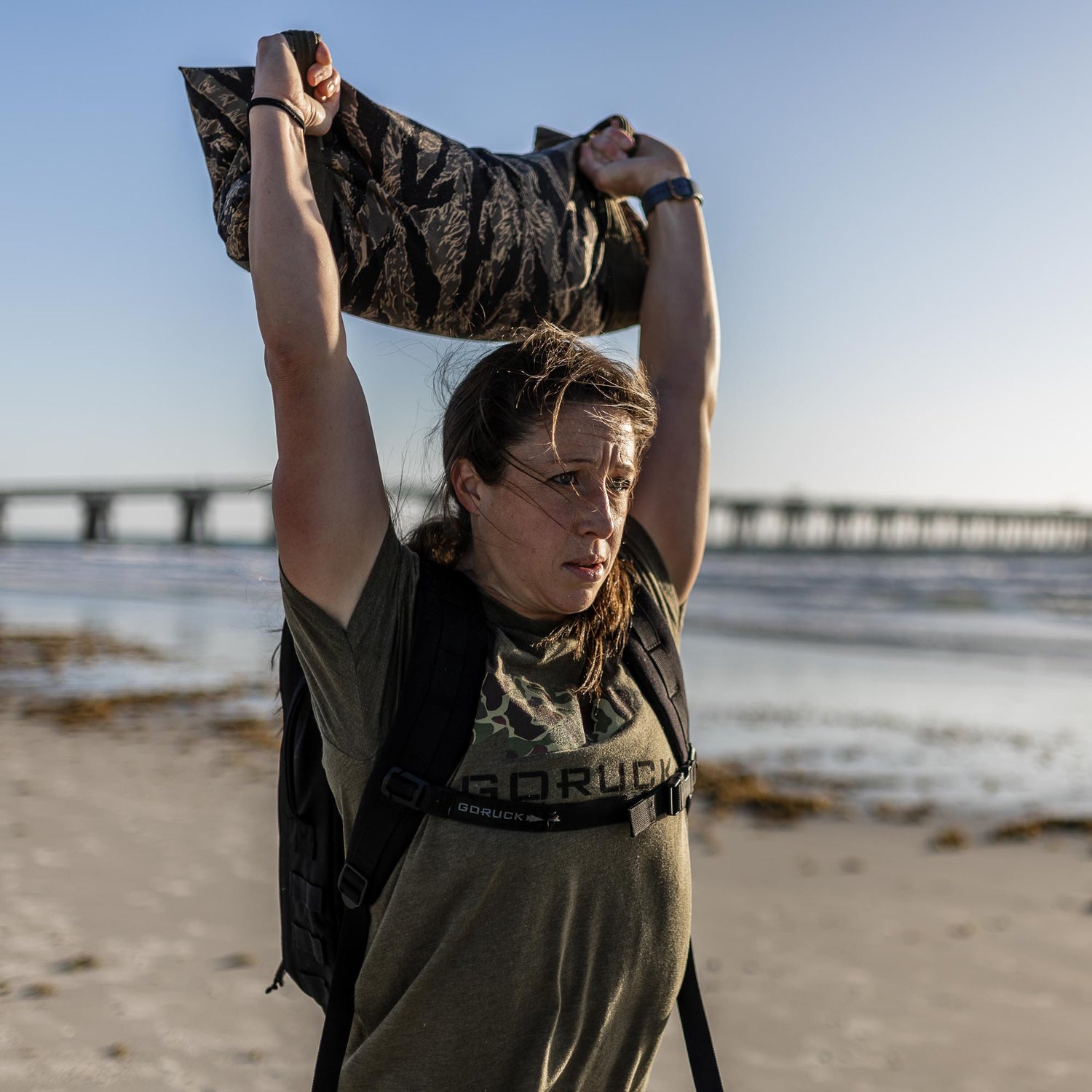 Someone lifts a Simple Training Sandbag overhead on a beach, with the pier and ocean visible in the background.