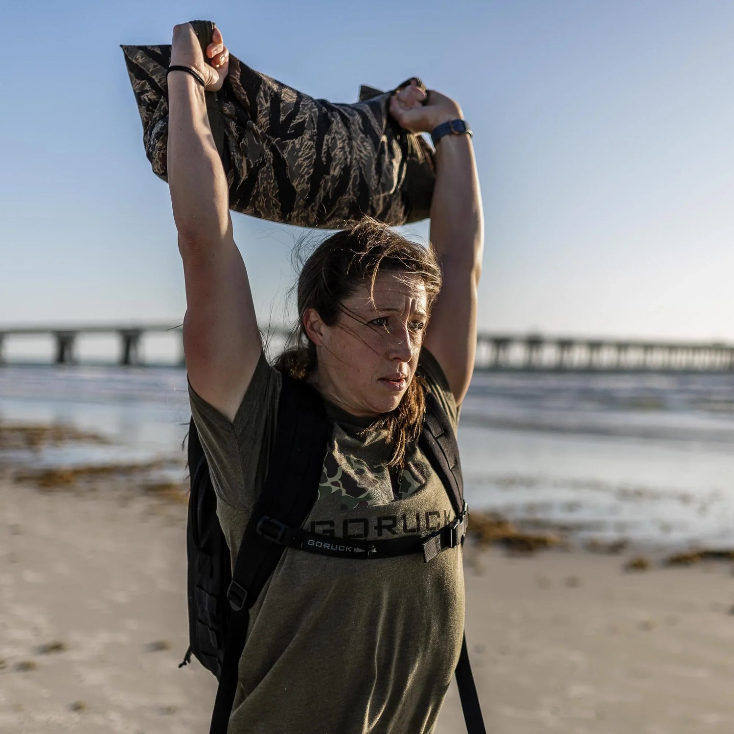 Woman lifting a weighted sandbag overhead on a beach wearing GORUCK gear with a bridge in the background