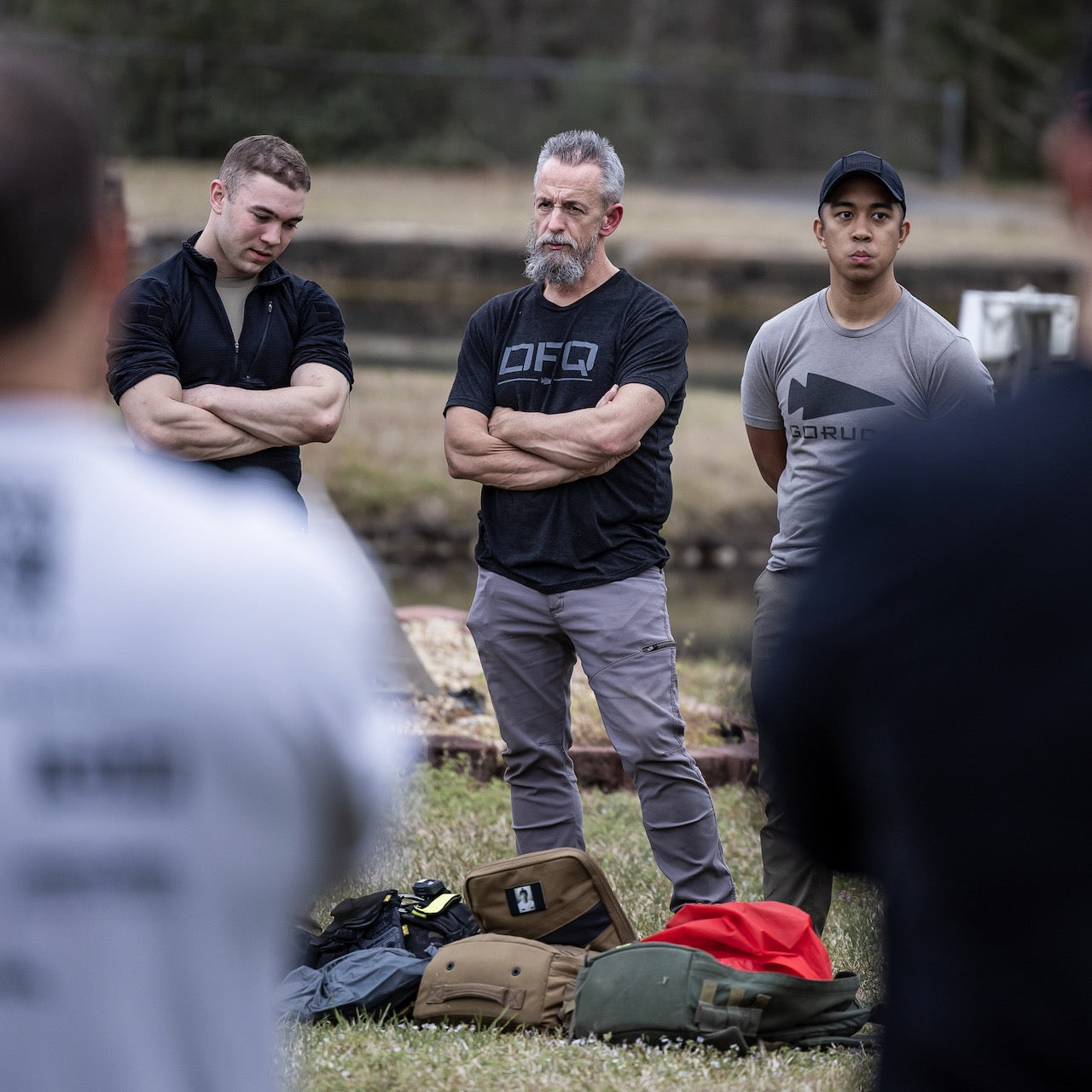 Three men stand outdoors with arms crossed, listening to someone off-frame. Each wears a DFQ Tee - Tri-BlendX, while their bags rest on the grass in front of them.