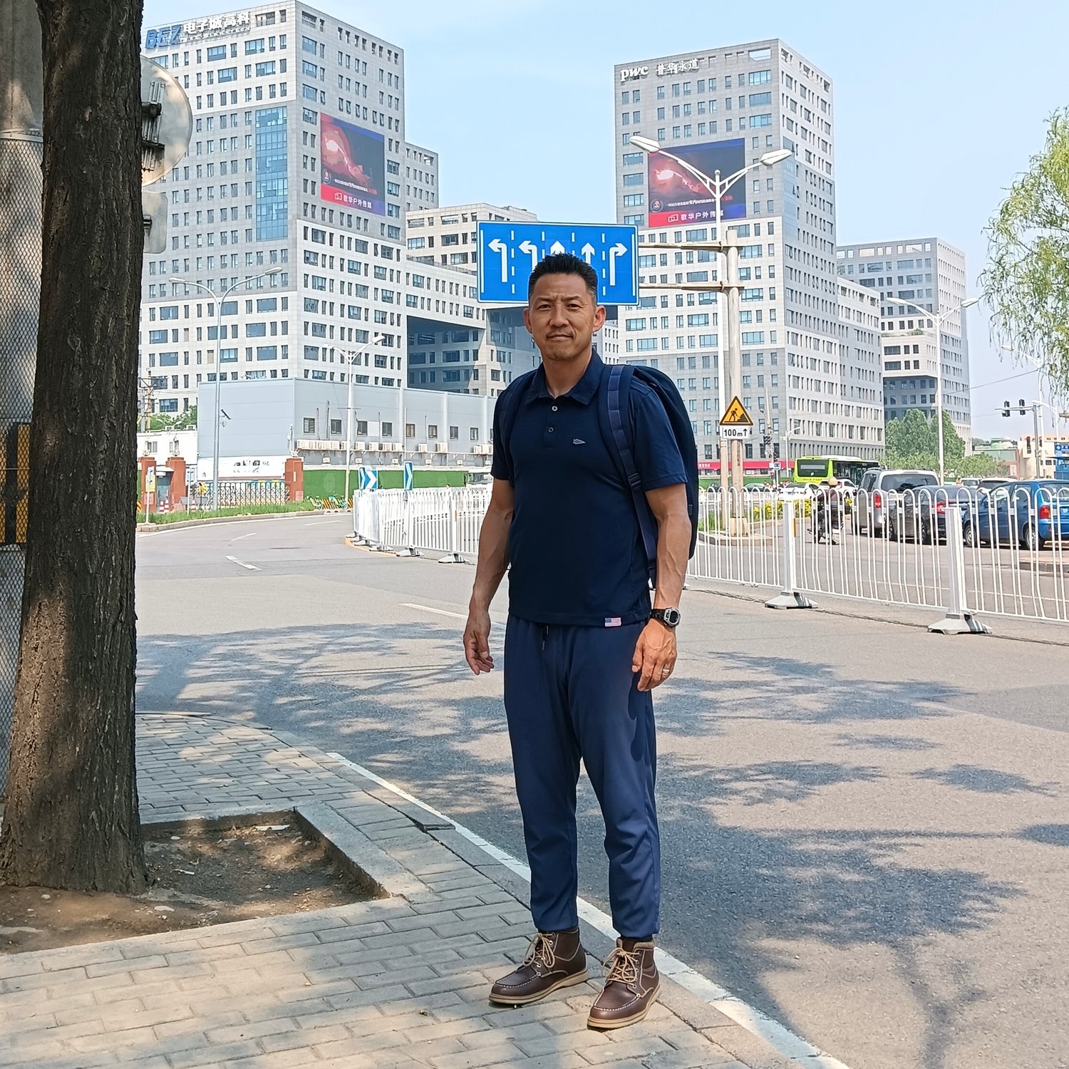 A man in blue clothes stands on a city sidewalk with tall buildings and parked cars in the background.