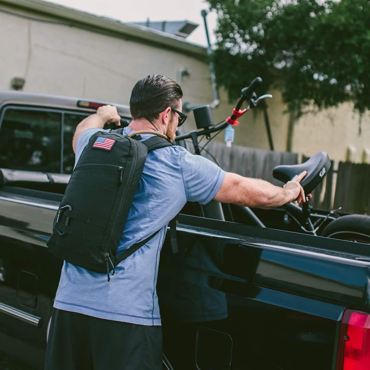 Man wearing GORUCK rucksack with USA flag patch loading bicycle into pickup truck outdoors