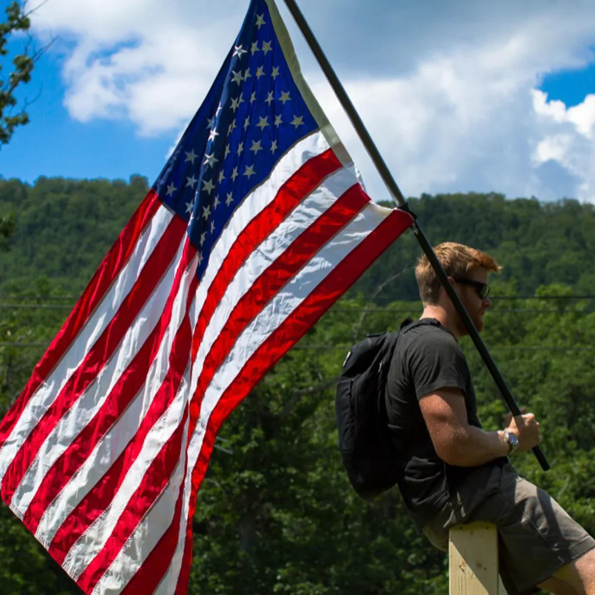 A man with a Bullet Ruck USA Throwback daypack sits on a post holding a large American flag, surrounded by trees and blue sky.