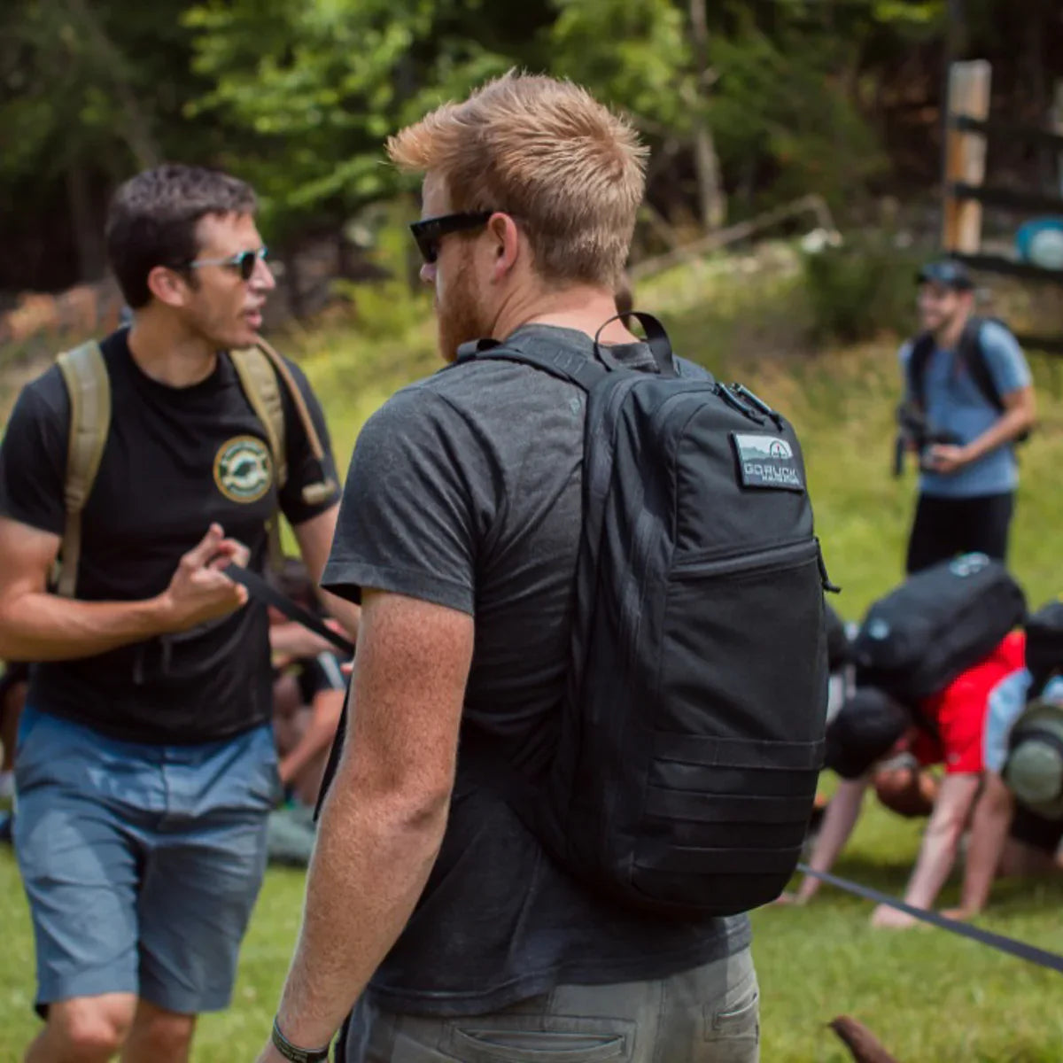 Man outdoors wearing a black GORUCK rucksack with group training in background on grassy field