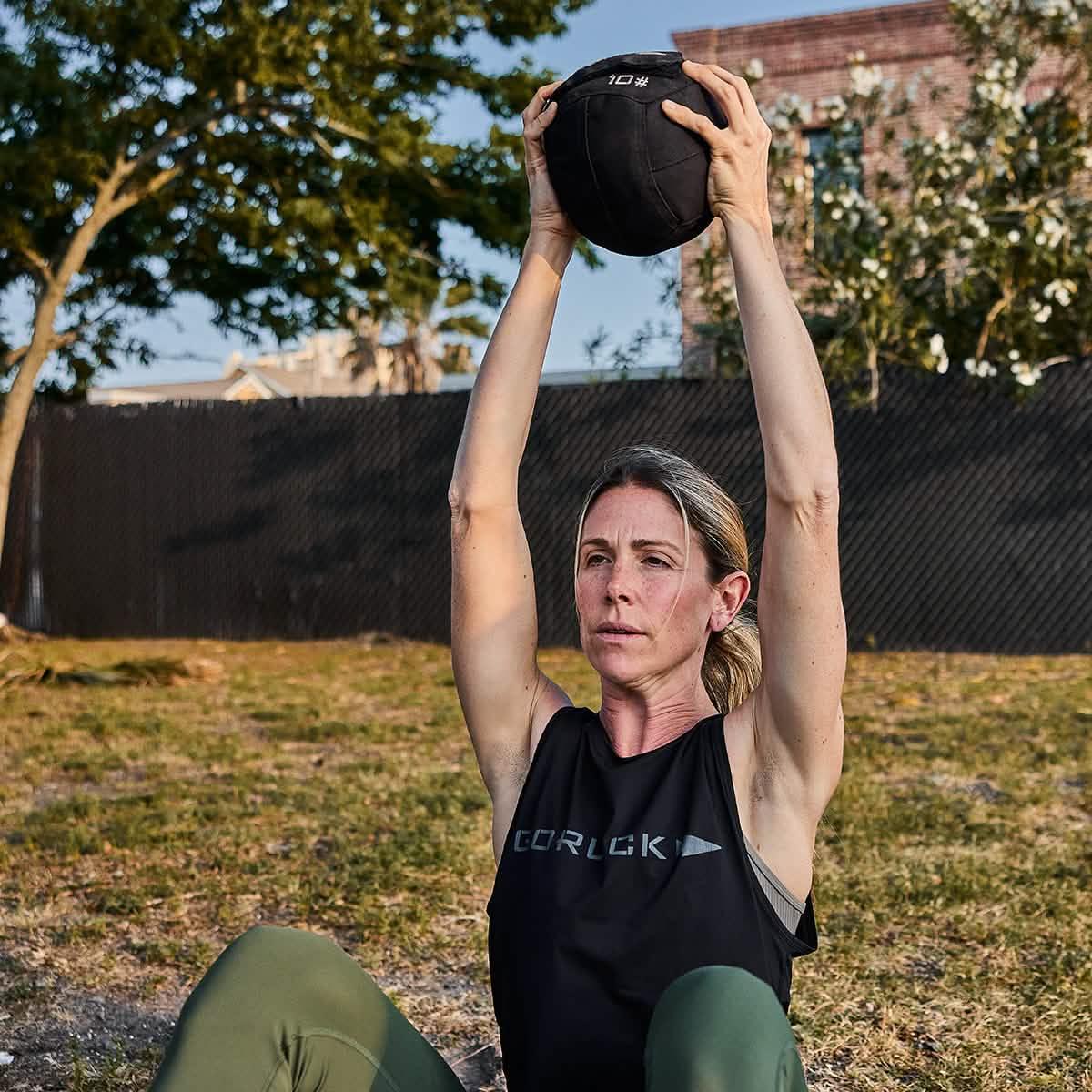 Woman exercising outdoors holding a 10 lb black GORUCK weighted ball overhead, wearing a black GORUCK tank top