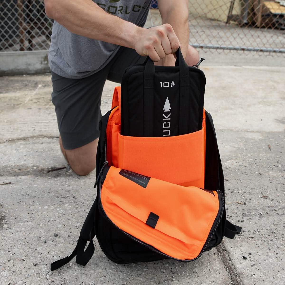 Person placing a black 10lb GORUCK weight plate into an open black and bright orange rucksack on pavement next to chain-link fence