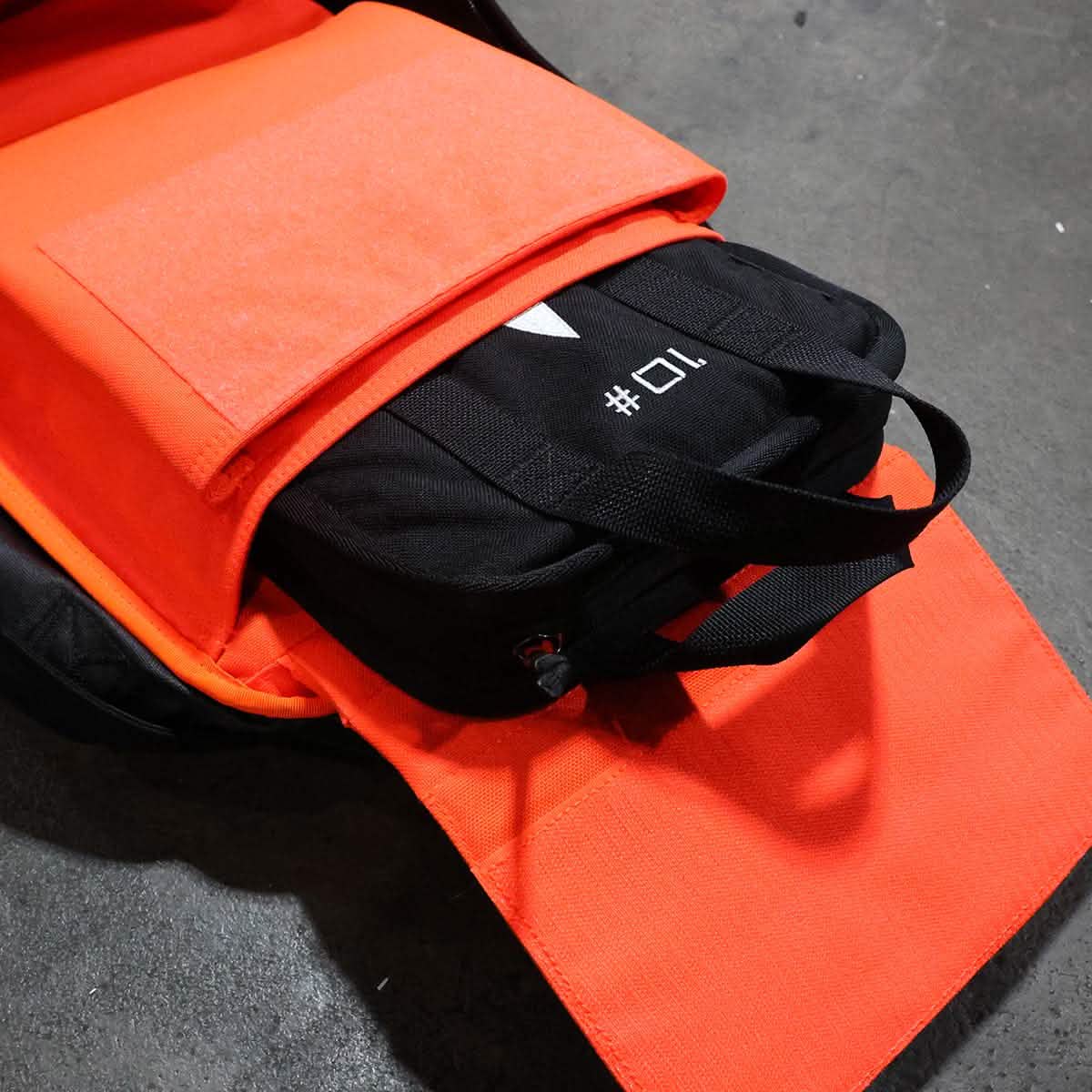 A Rucker backpack in orange lies open on a dark surface, displaying a neatly packed black item marked with 01. This sturdy pack, featuring GORUCK's Lifetime Guarantee, is ideal for carrying essentials like the durable Sand Ruck Plates.