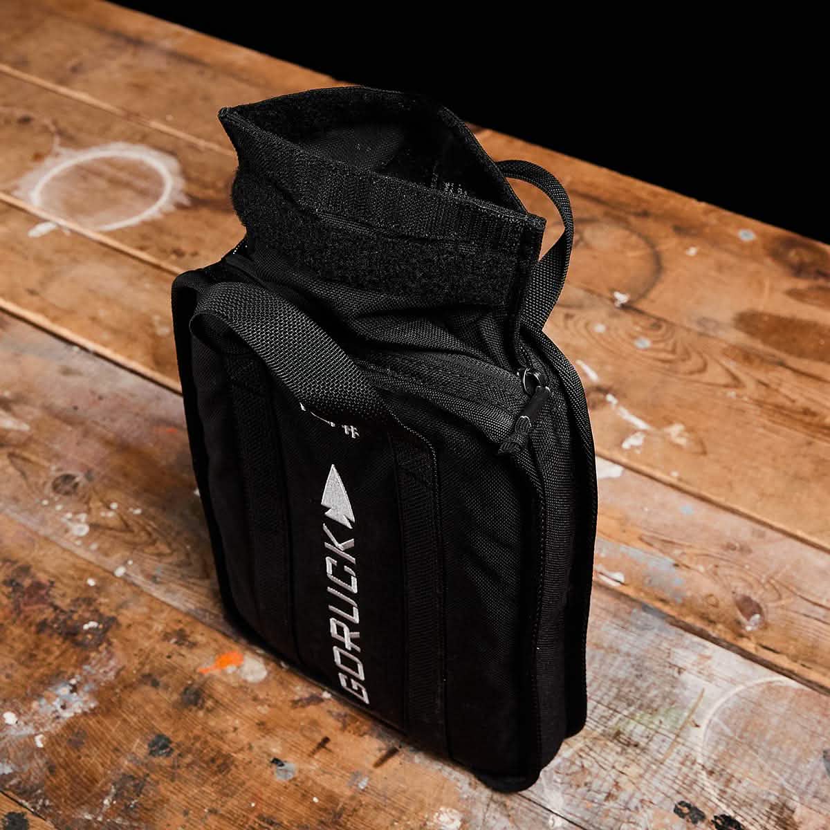 A black GORUCK Sand Ruck Plate bag sits upright on a worn wooden surface. Its top is open, revealing part of the strap and zipper beside a rugged Ruck Plate. The bag features a white logo with an arrow and comes with a lifetime guarantee.