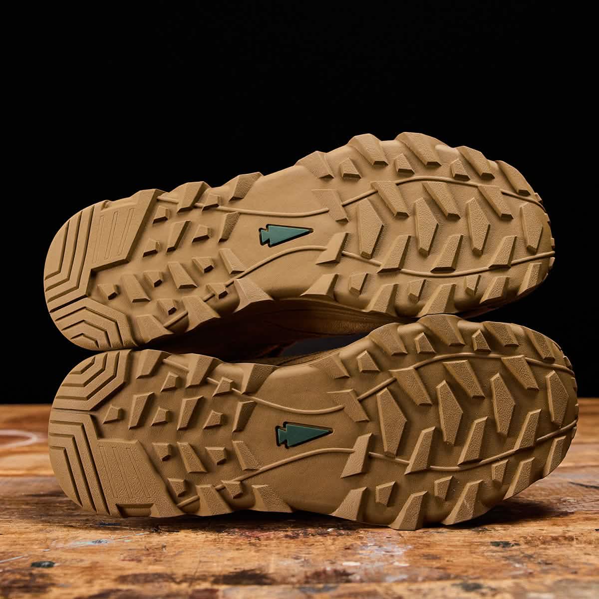 The image showcases the soles of the GORUCK MACV-2 Safety Boot - Mid Top, featuring rugged and textured tread patterns set against a dark background. These work boots are designed with angular details and sport green triangular logos near the center, adhering to Special Forces Standards. They are placed neatly on a wooden surface.