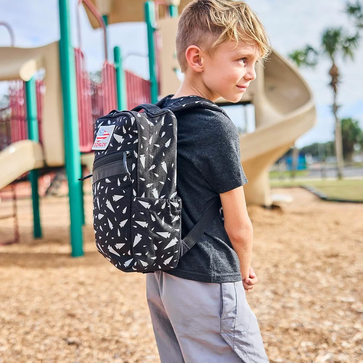A young boy stands near a playground slide, looking to the side, wearing the REFLECTIVE KR1 durable book bag.