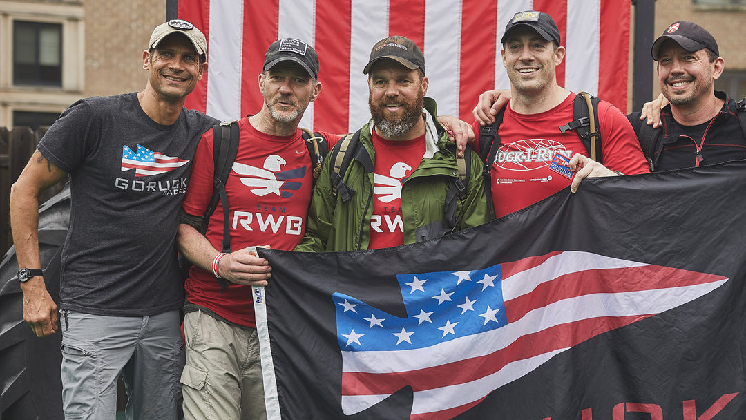 Five men smiling in front of an American flag, holding a black flag with a stylized US flag design.