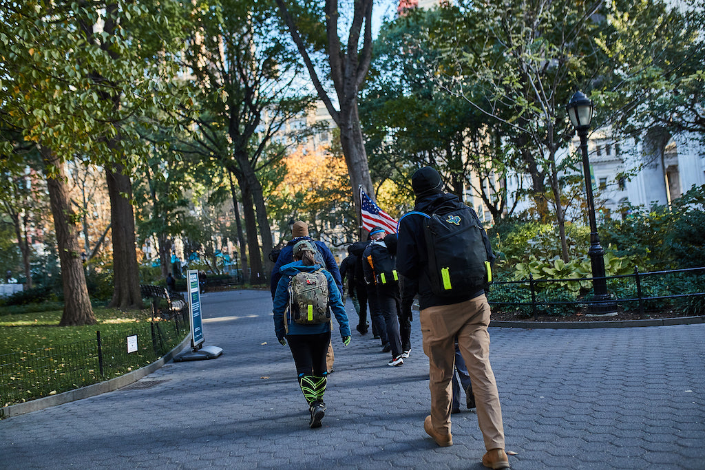 A group of people with backpacks walk through a city park, one carrying an American flag.