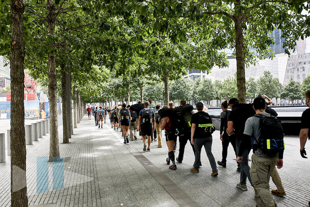 A group of people with backpacks walk under trees along a city sidewalk on a sunny day.