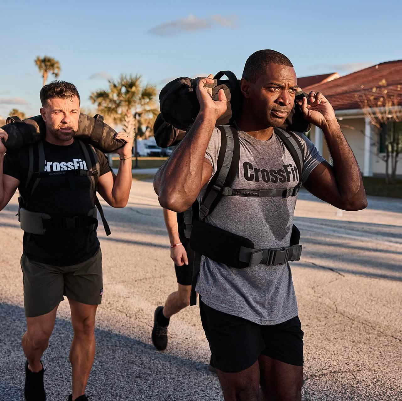 Two men wearing CrossFit shirts carrying GORUCK weighted bags outdoors during a fitness rucking workout