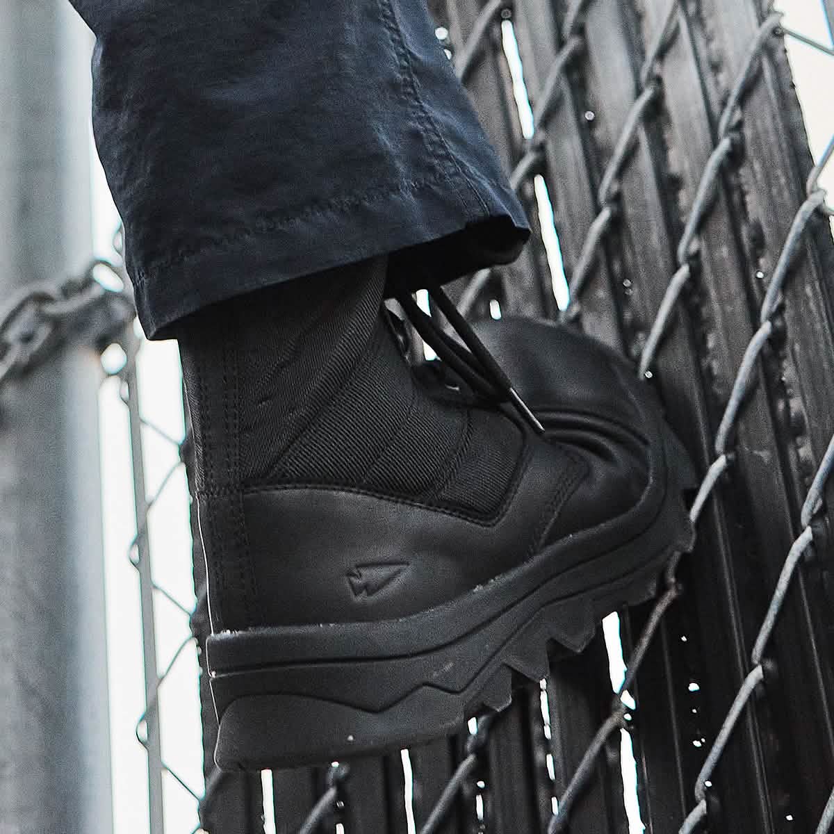 A close-up of a MACV-2 Safety Boot - High Top by GORUCK, designed to meet Special Forces standards, stepping on a chain-link fence. The black boot showcases thick, rugged soles and is worn with dark pants. The angled metallic fence suggests an outdoor or urban environment.