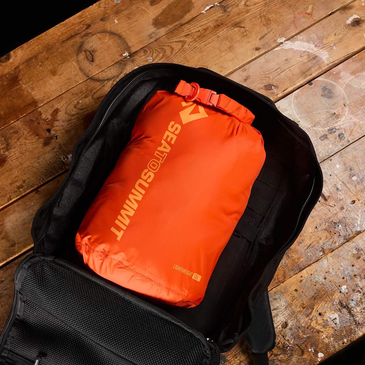 A black backpack on a wooden surface holds an orange Sea to Summit Dry Sack featuring a waterproof closure, made from lightweight nylon.
