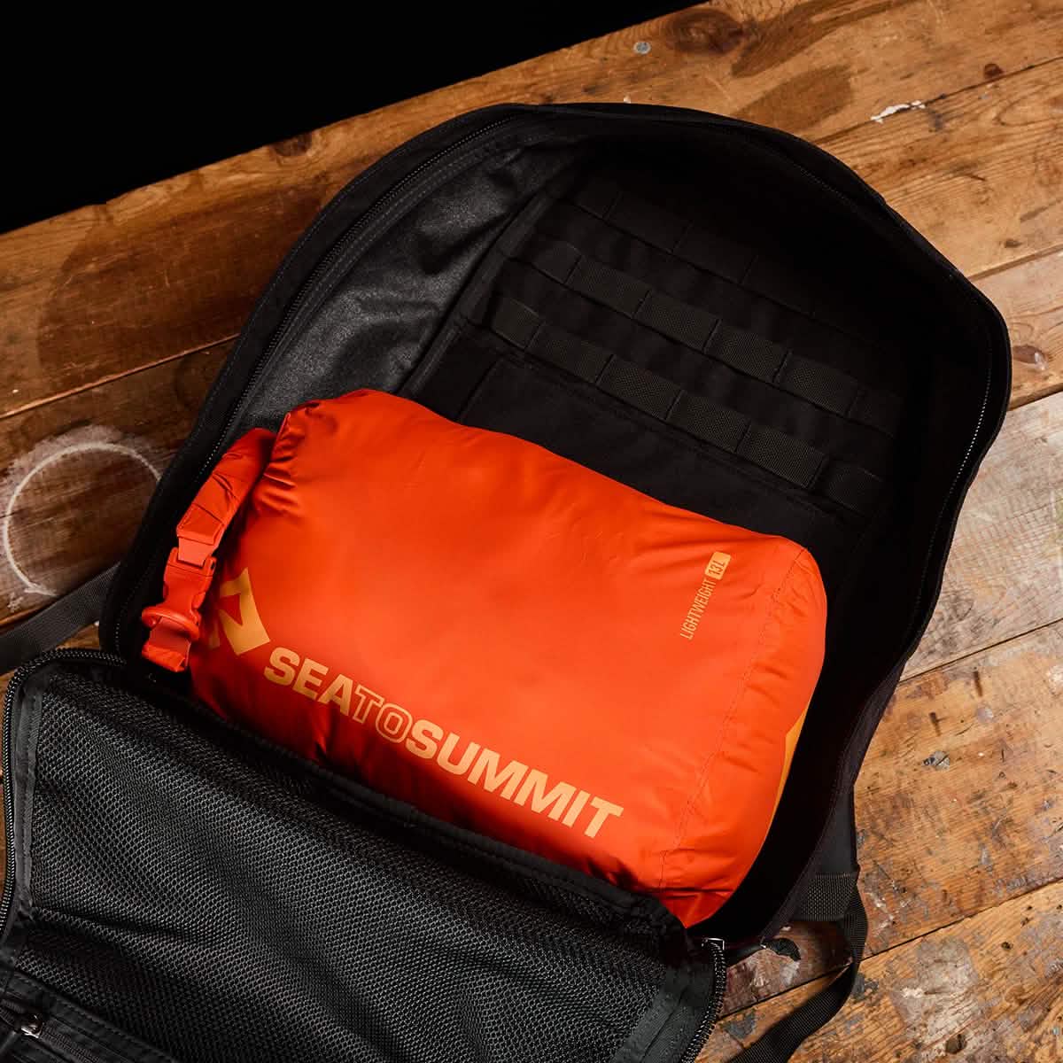 A black backpack sits on a wooden surface with an orange Sea to Summit Dry Sack neatly packed inside. The backpack is open, displaying the Dry Sack occupying the main compartment, protected by its lightweight nylon exterior and waterproof closure.