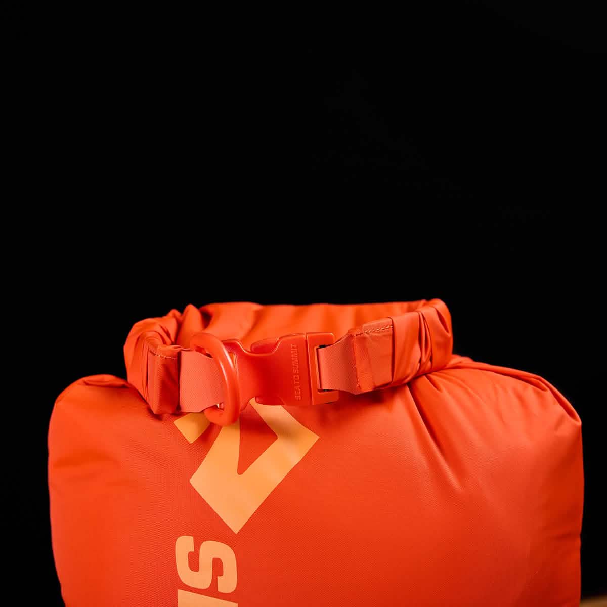 In the foreground of a black background is the Sea to Summit Dry Sack, an orange roll-top backpack made from lightweight nylon featuring a waterproof closure and buckle. It showcases a striking, stylized yellow logo.